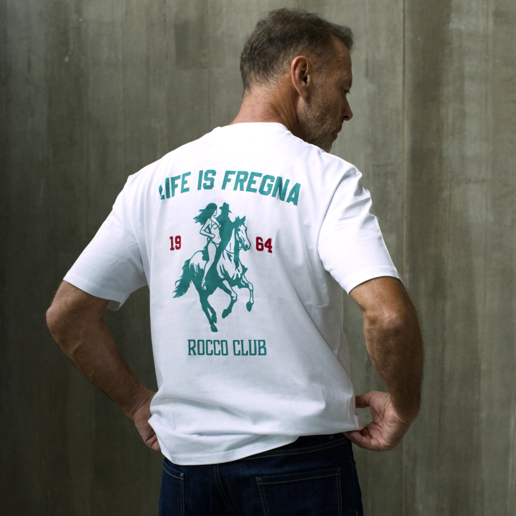 A man wearing a white t-shirt with a green and red graphic print on the back, standing against a textured gray concrete wall.