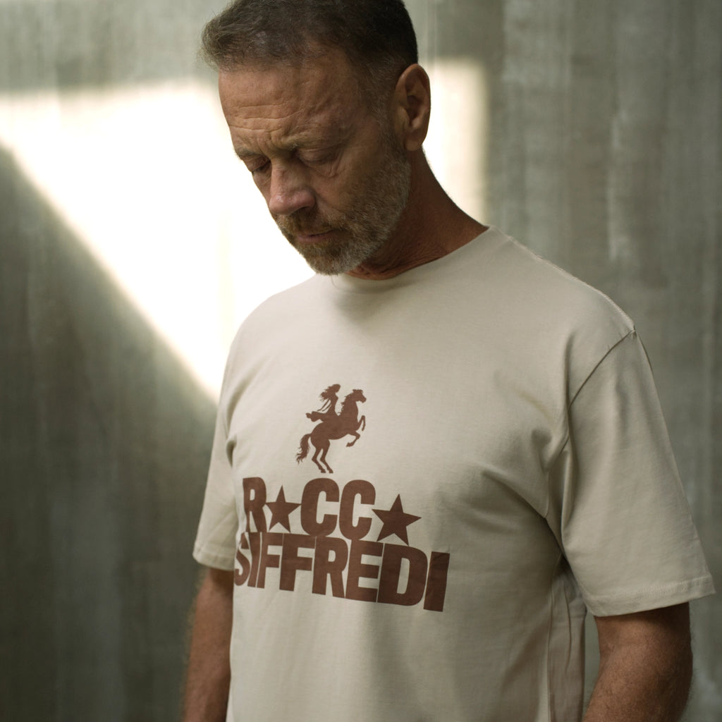 A man wearing a beige t-shirt with a brown graphic print that reads 'ROCCO SIFFREDI' with a rider on a horse silhouette, photographed against a textured gray wall with dramatic lighting.